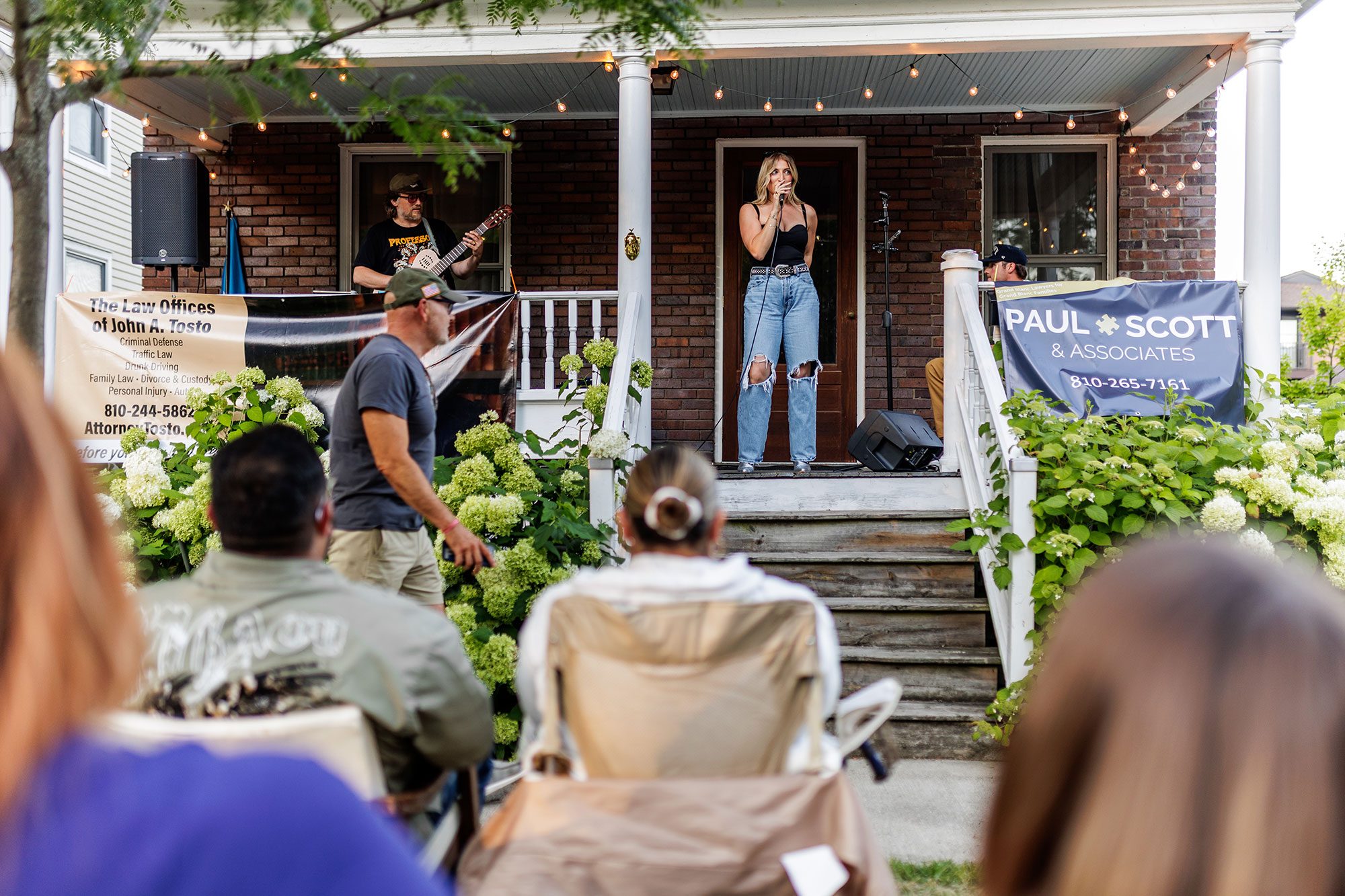 Singer performing on a brick porch with string lights, audience watching from lawn chairs